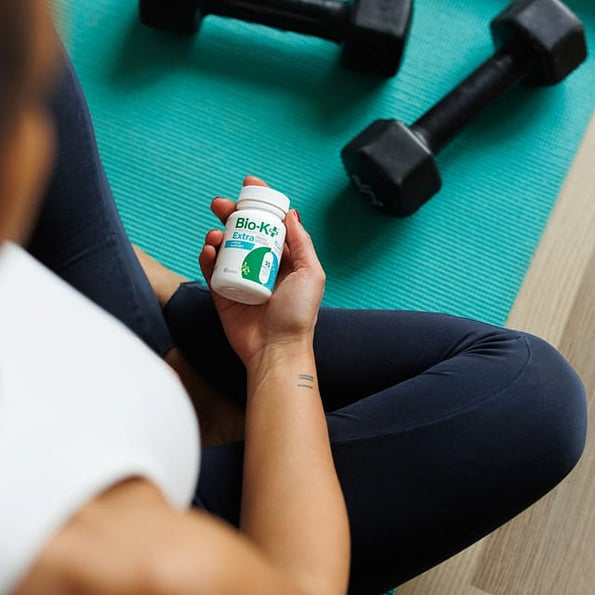 Person sitting on gym floor holding Bio-K+ probiotic bottle with dumbbells, supporting stress recovery after exercise.